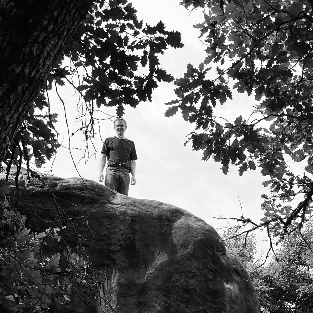 Tom de Gay standing on top of a large boulder in the forest of Fontainebleau, framed by oak branches.